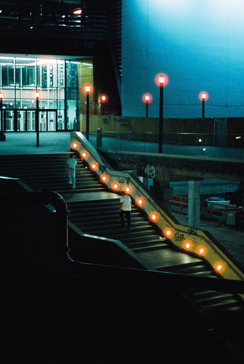Two men walk up a wide outdoor staircase with orange lights embedded in the handrail at night.