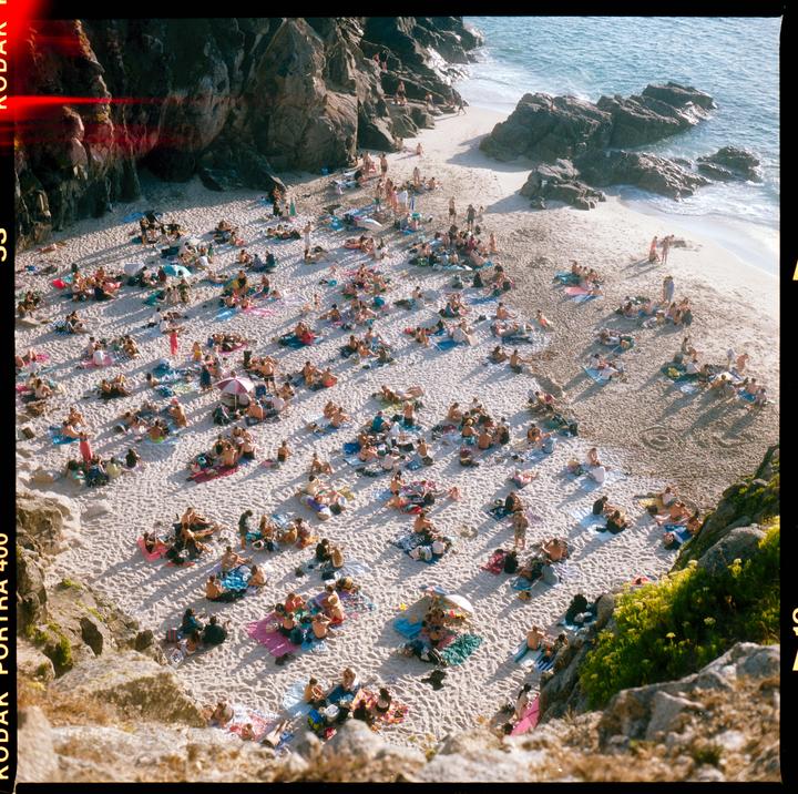 A large crowd of people are sunbathing on a sandy beach next to a rocky coastline.