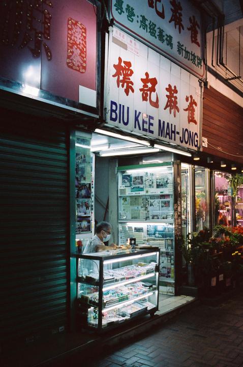 A man wearing a face mask sits behind a counter in a brightly lit storefront at night.