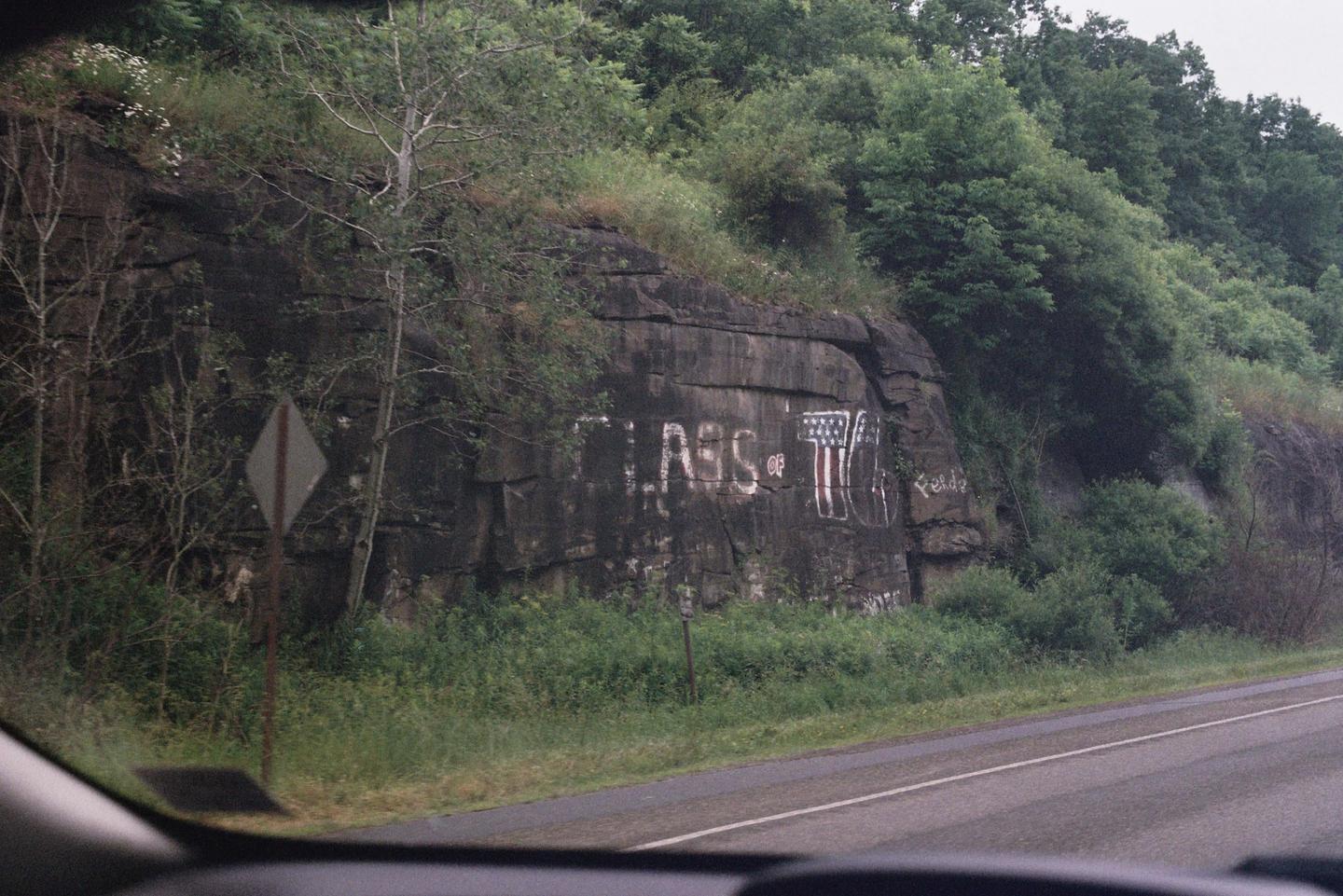 A rock face with "CLASS of 76" painted on it is visible from a car on a road.