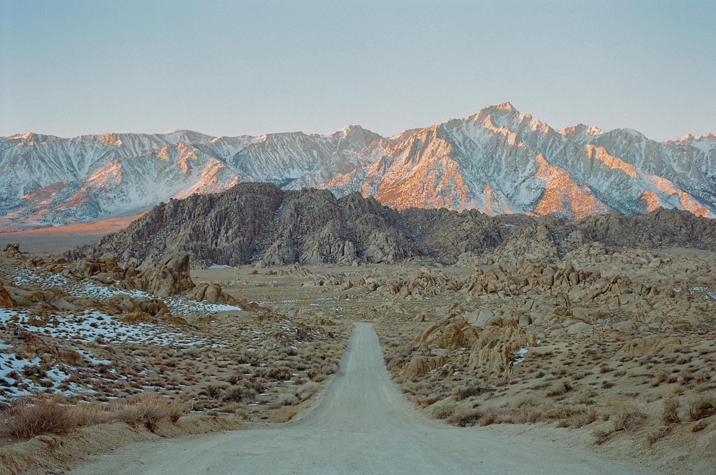 A dirt road leads toward snow-capped mountains with rocky hills in the foreground.
