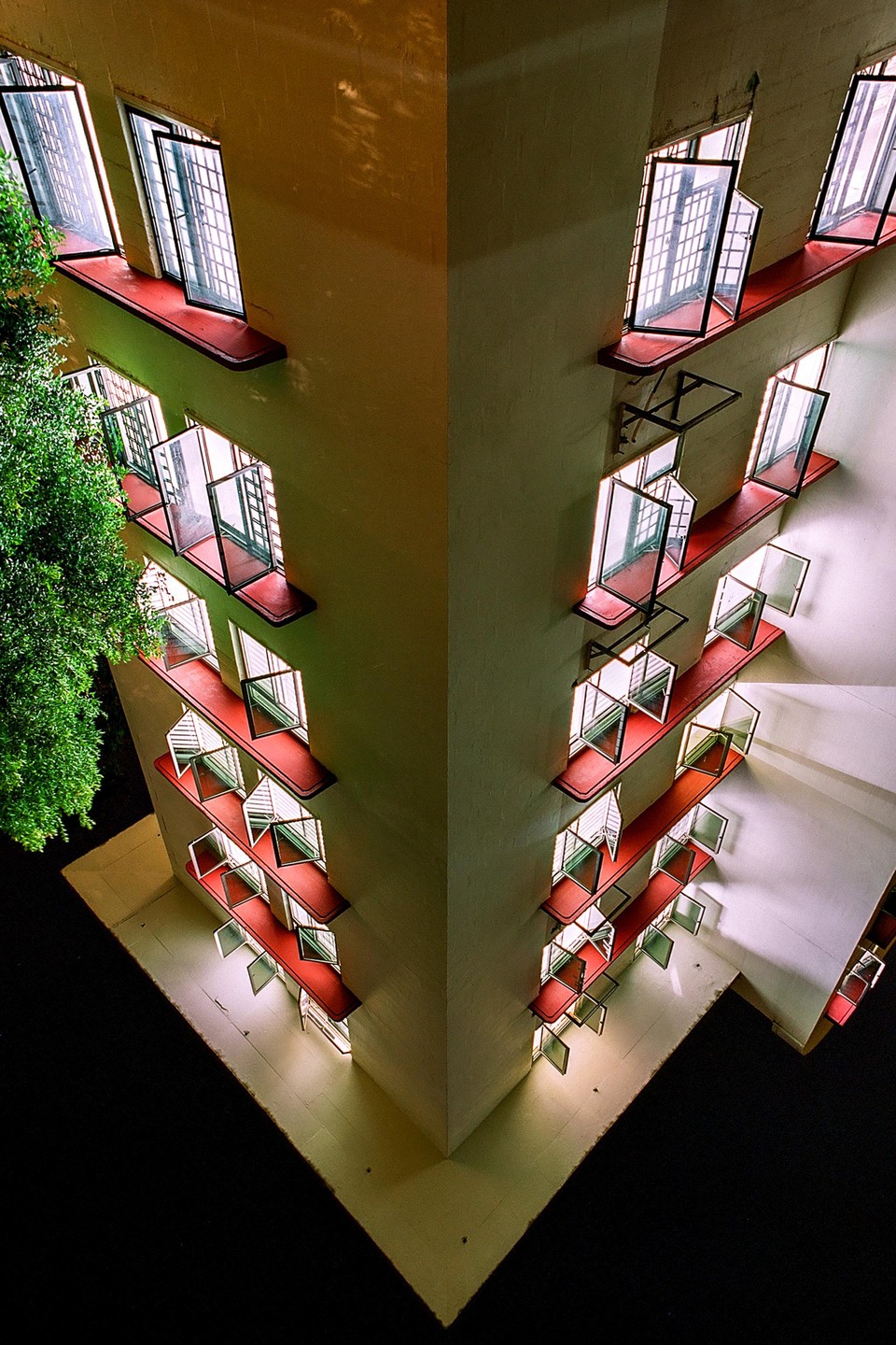 A tall building with many open windows and red ledges is illuminated at night, with a tree visible on the left.