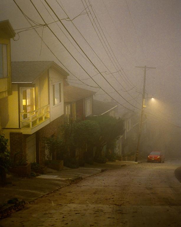 A red car is parked on a foggy street lined with houses and power lines.