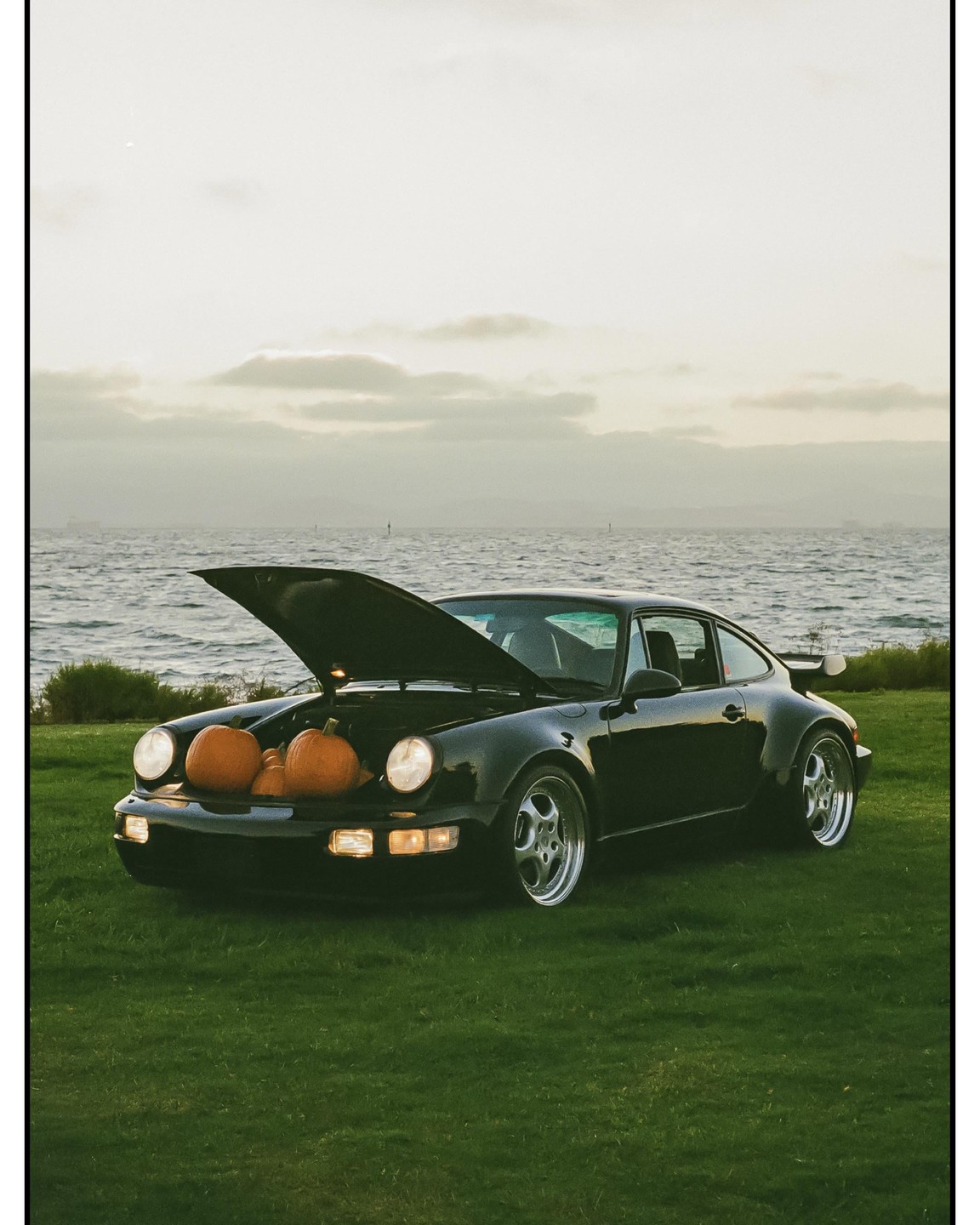 A black sports car with its hood open, revealing several pumpkins inside, is parked on a grassy area beside a body of water under a cloudy sky.