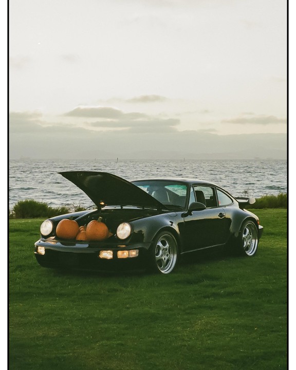 A black sports car with its hood open, revealing several pumpkins inside, is parked on a grassy area beside a body of water under a cloudy sky.