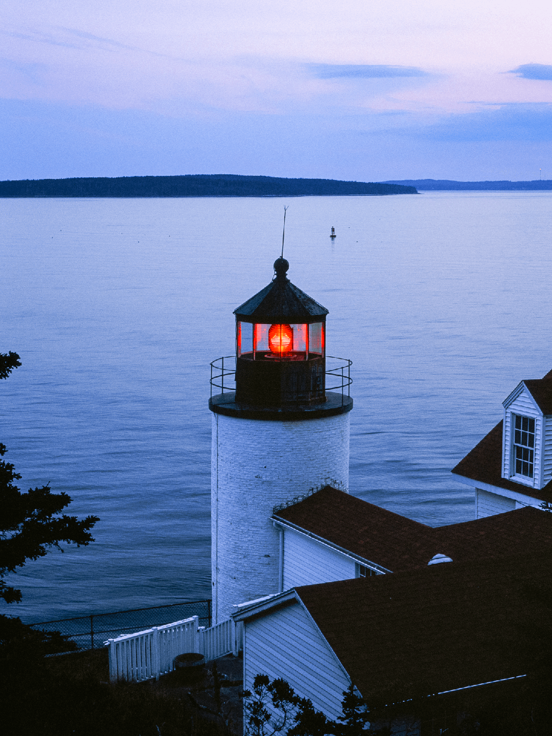 A lighthouse with a glowing red light stands by the ocean at dusk.