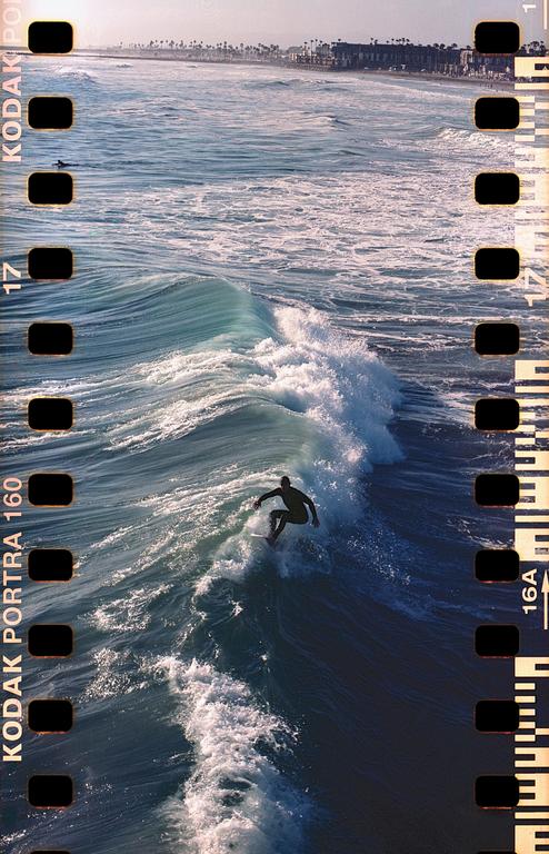 A surfer rides a wave with a town visible in the distance along the coast.