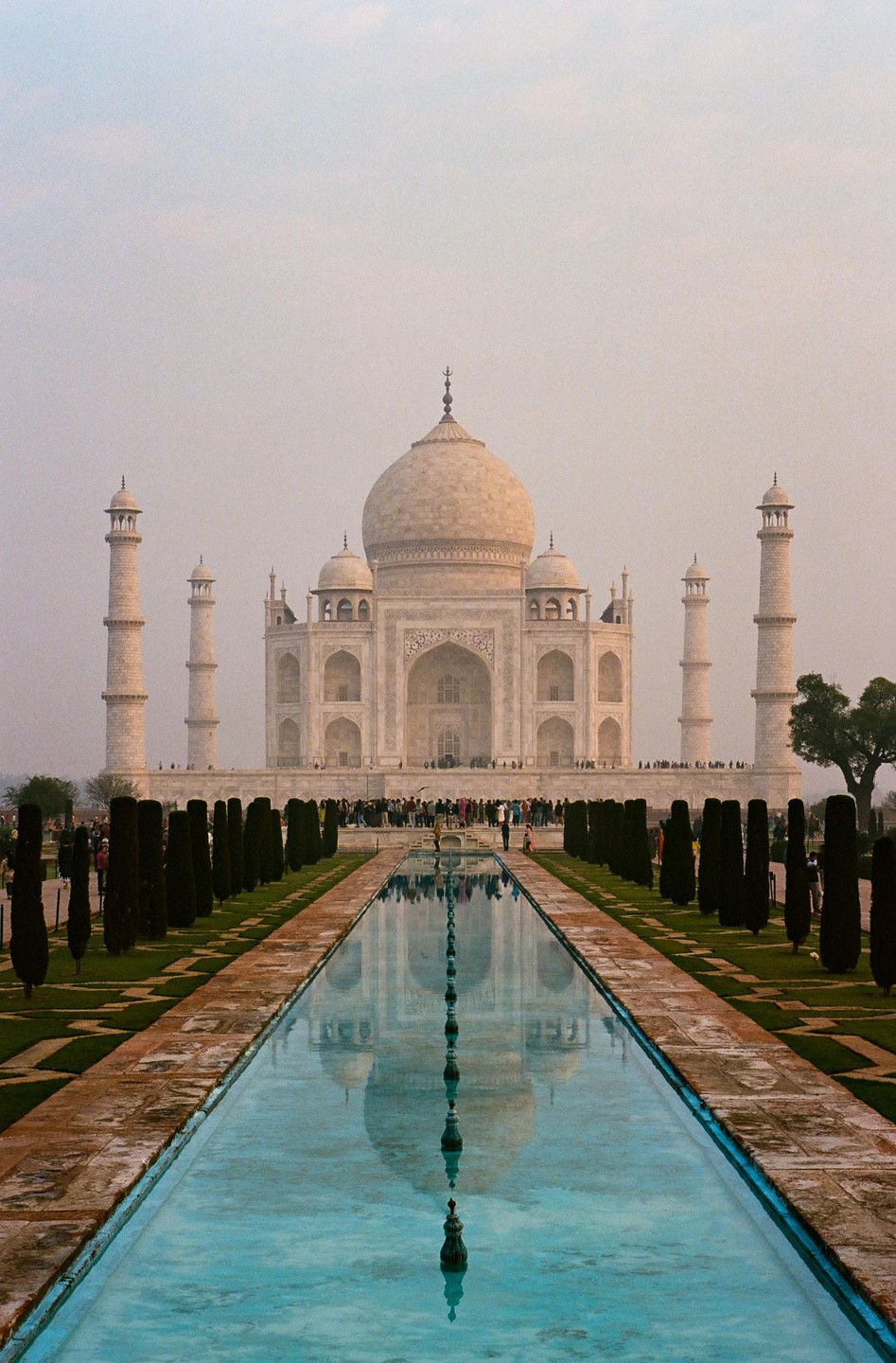 The Taj Mahal is reflected in a long, narrow pool of water, with people gathered around the monument.