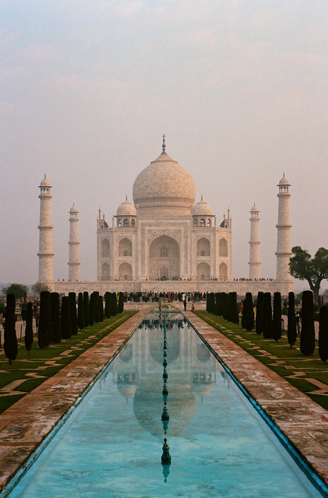 The Taj Mahal is reflected in a long, narrow pool of water, with people gathered around the monument.