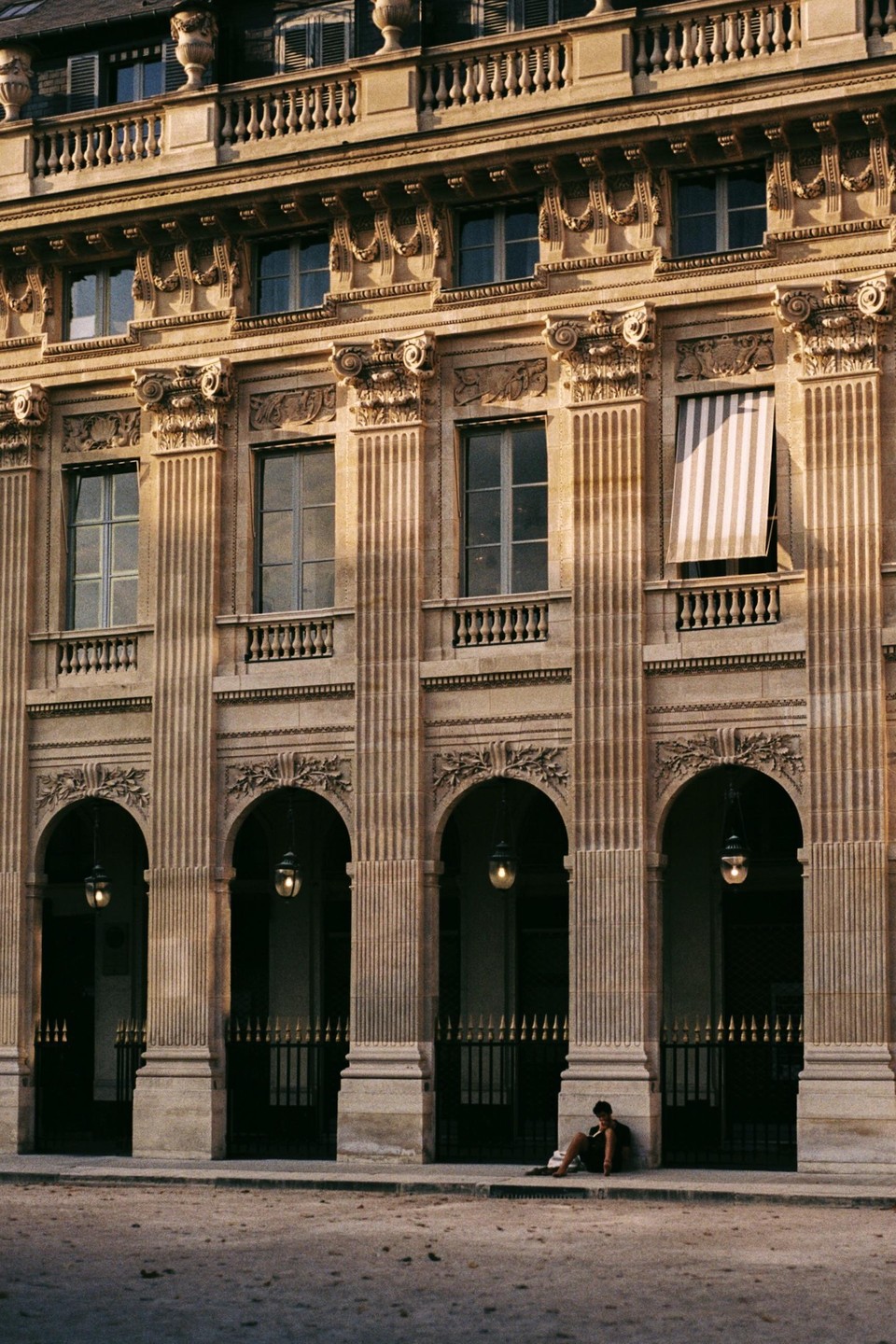 A person sits on the ground in front of a large building with arches and columns.