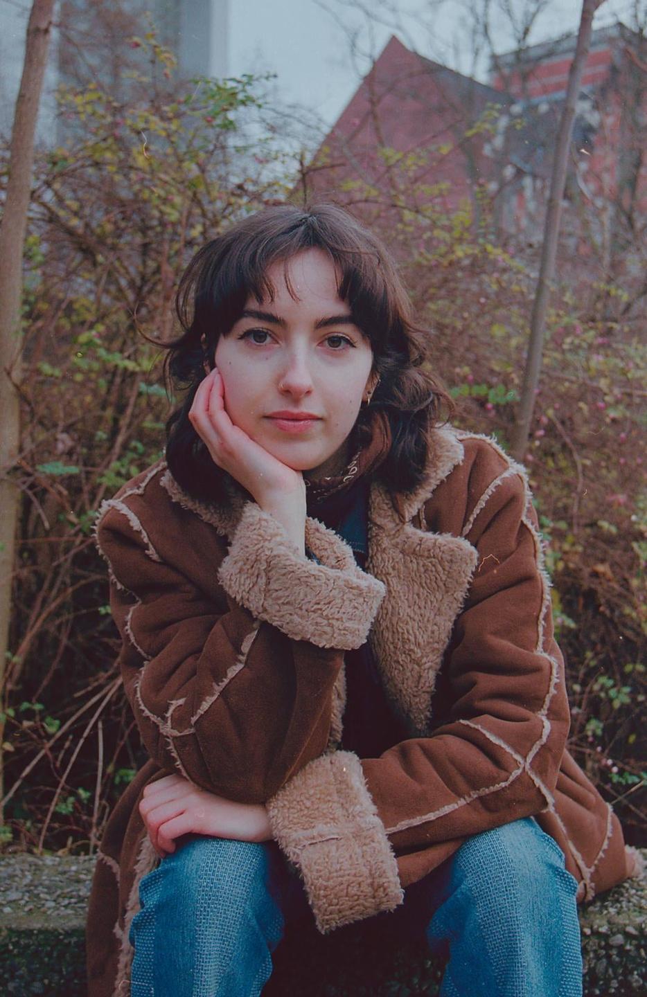 A woman wearing a brown coat with a fur collar and blue jeans sits outdoors with her chin resting on her hand.