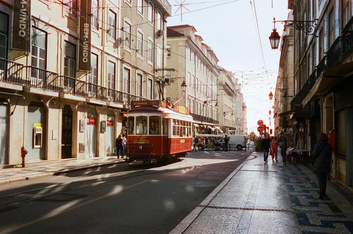 A red tram is parked on a street lined with buildings and people walking on the sidewalk.