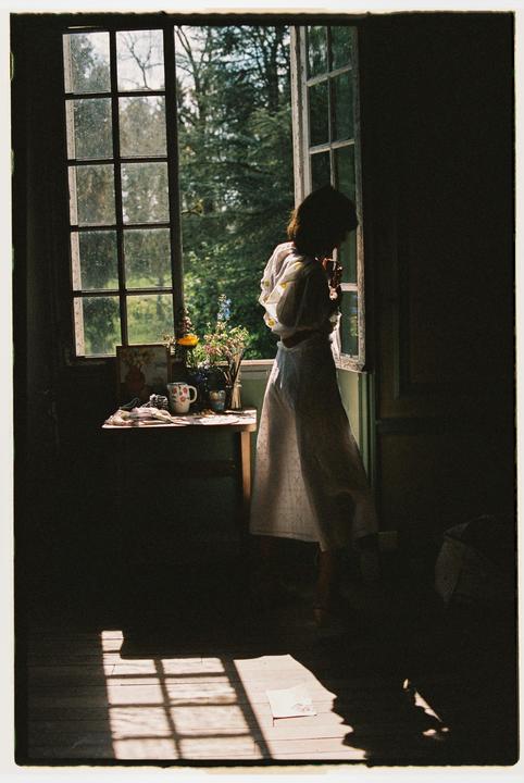 A woman stands by an open window, looking out at a lush green landscape, with sunlight streaming into the room.