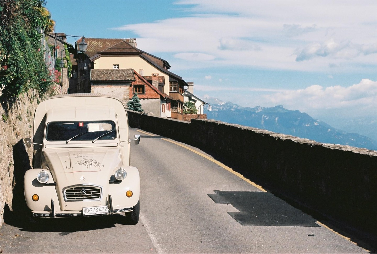 A beige Citroen 2CV van is parked on a narrow road next to a stone wall, with buildings and mountains in the background.