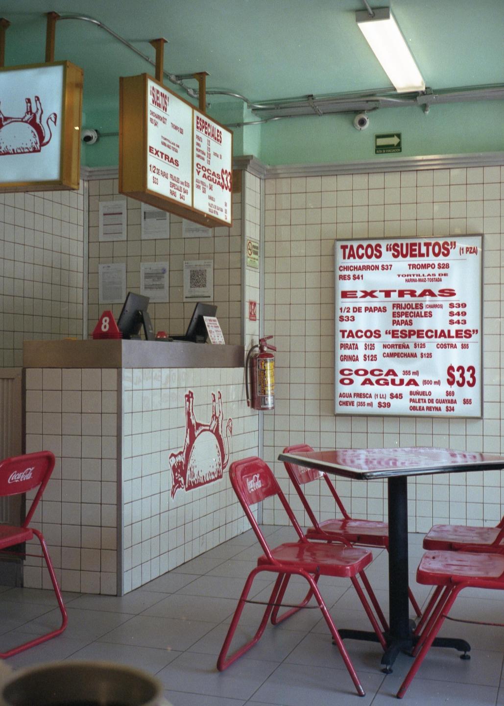 A tiled taqueria interior features red chairs and a table, with a menu board displaying various taco options.