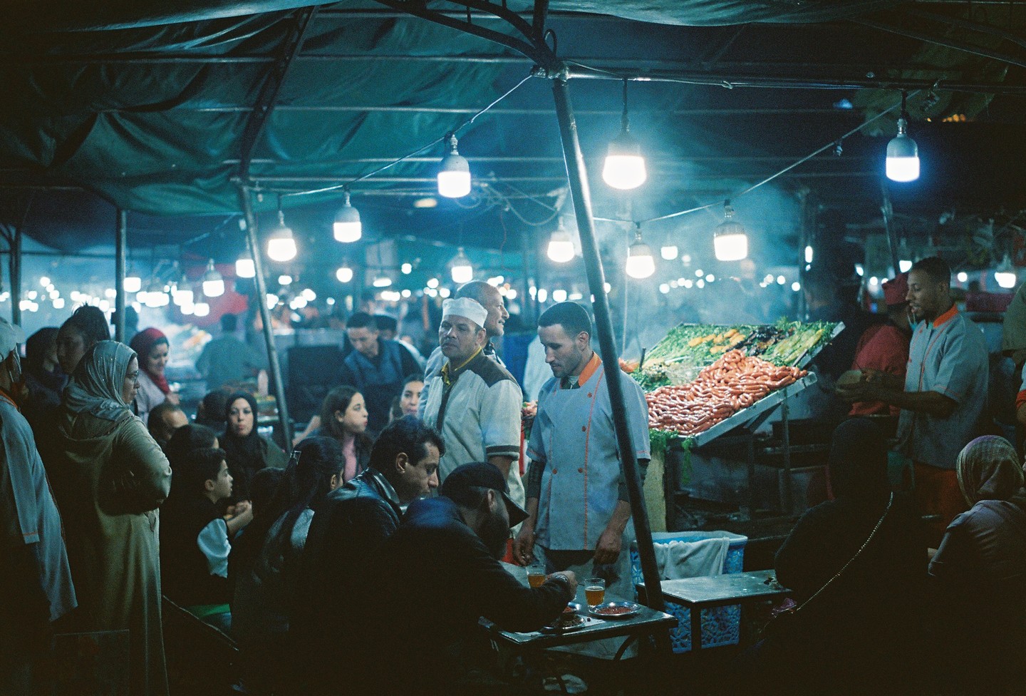 People gather under a canopy lit by hanging bulbs at a night market with food stalls.