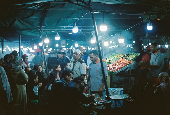 People gather under a canopy lit by hanging bulbs at a night market with food stalls.