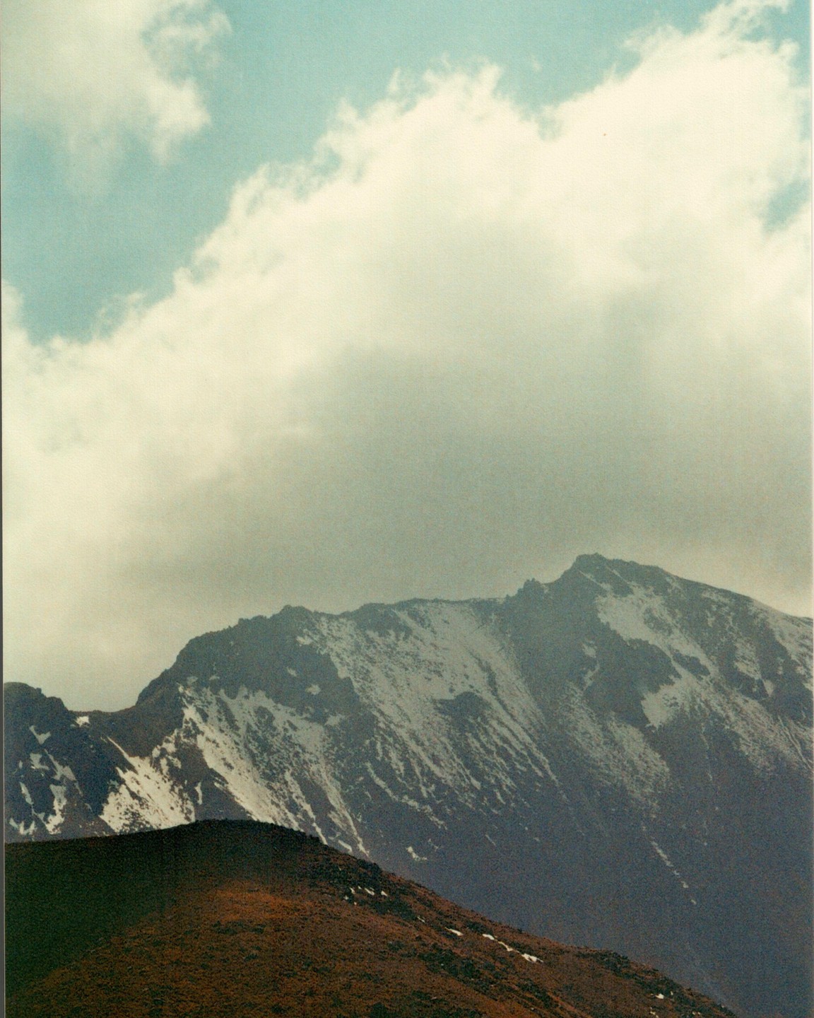 A snow-capped mountain peak rises above a brown hill under a cloudy sky.