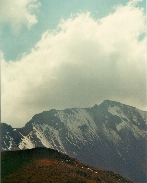 A snow-capped mountain peak rises above a brown hill under a cloudy sky.