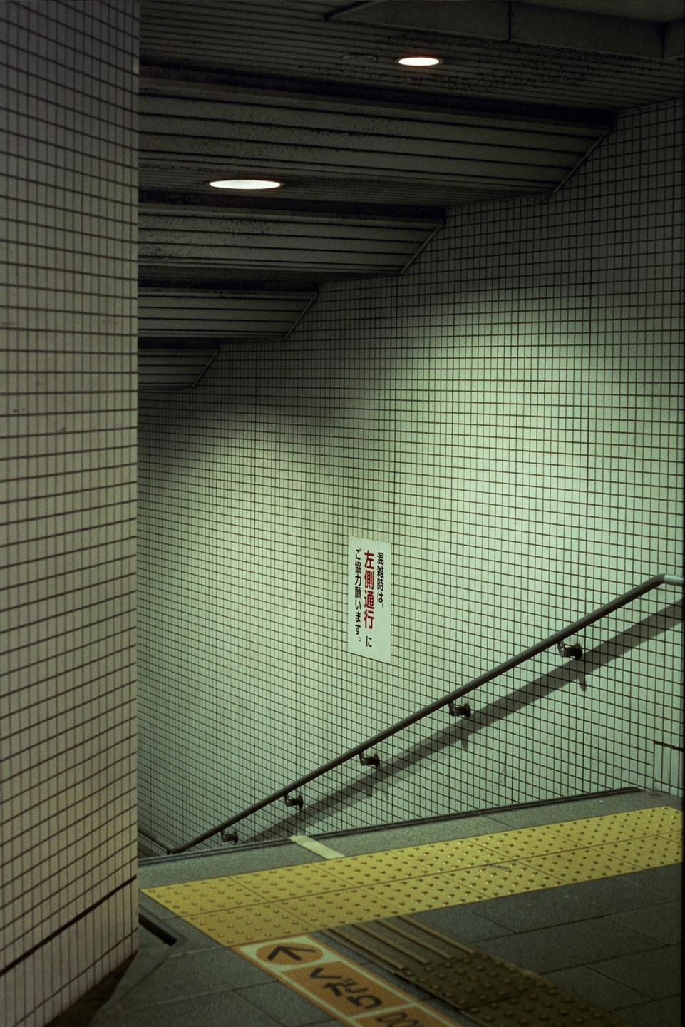 A tiled underground walkway with a handrail and a yellow tactile paving strip on the floor leads down a set of stairs.