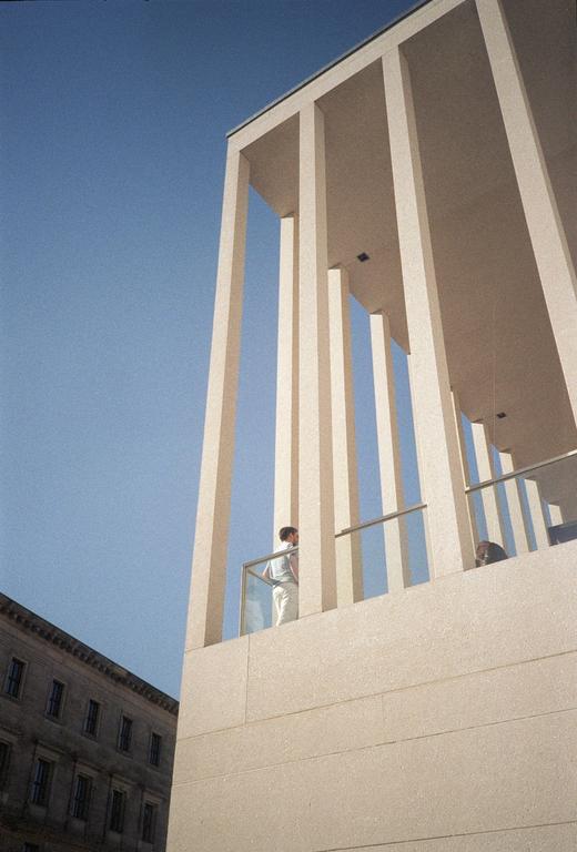 A person stands on a balcony of a modern building with tall pillars, looking out at another building under a clear blue sky.