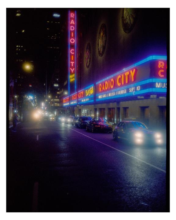 A street at night is illuminated by the neon signs of Radio City Music Hall and the headlights of cars.