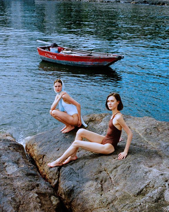 Two women in swimsuits pose on rocks by the water with a small red boat in the background.