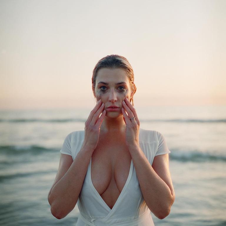 A woman with smudged makeup on her face stands in front of the ocean with her hands on her cheeks.
