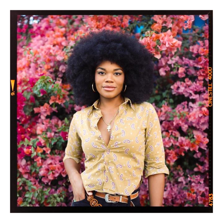 A woman with an afro stands in front of a vibrant pink and red floral bush.