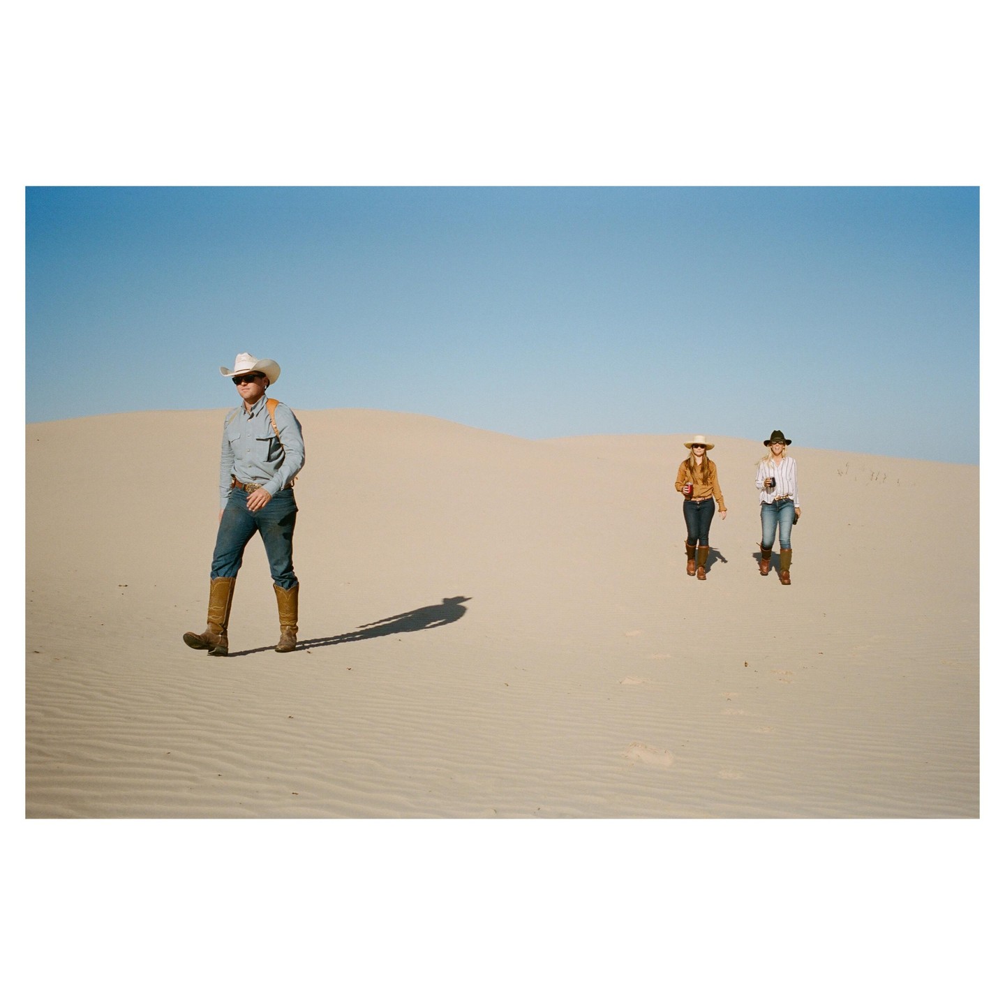 A man and two women wearing cowboy hats and boots walk across a desert landscape with sand dunes.