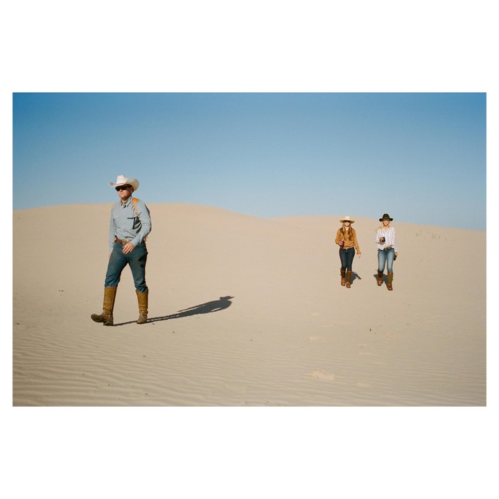 A man and two women wearing cowboy hats and boots walk across a desert landscape with sand dunes.