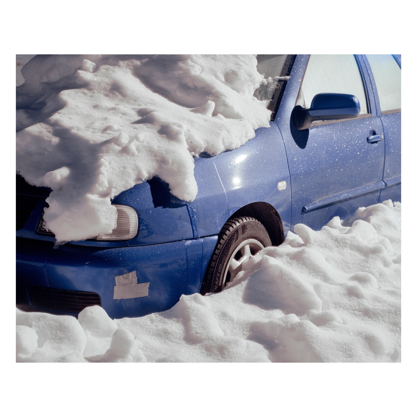 A blue car is partially buried in deep snow on a sunny day.
