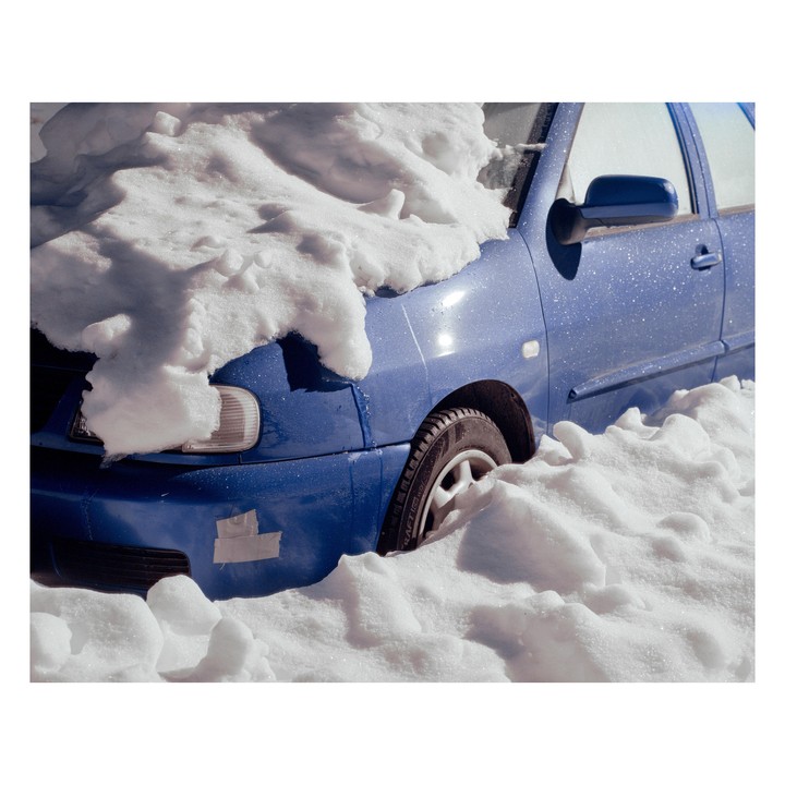 A blue car is partially buried in deep snow on a sunny day.