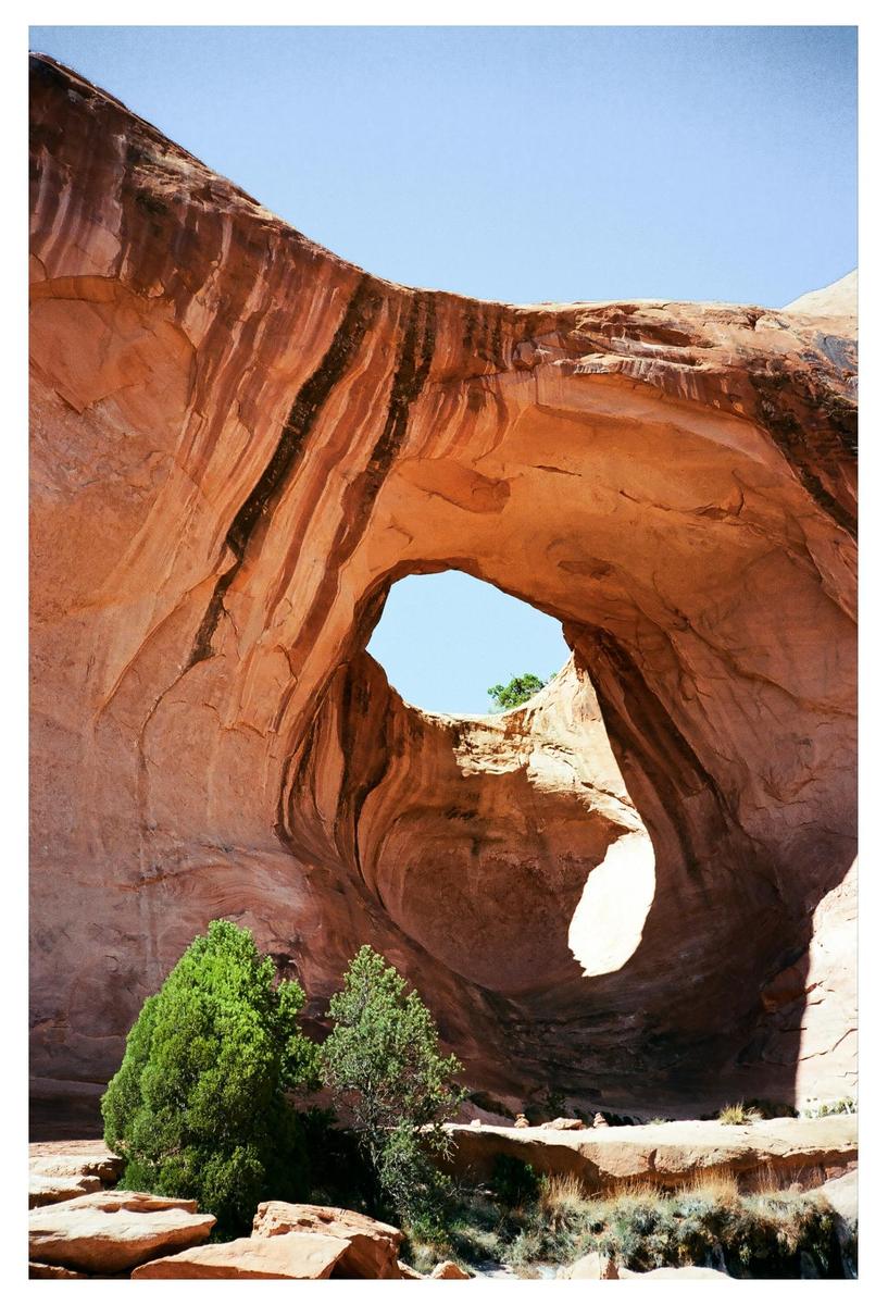 A large rock formation with a natural arch is visible under a clear sky, with green trees at its base.