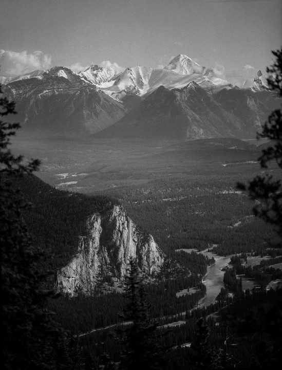 A river winds through a valley with a large rock formation and snow-capped mountains in the distance.