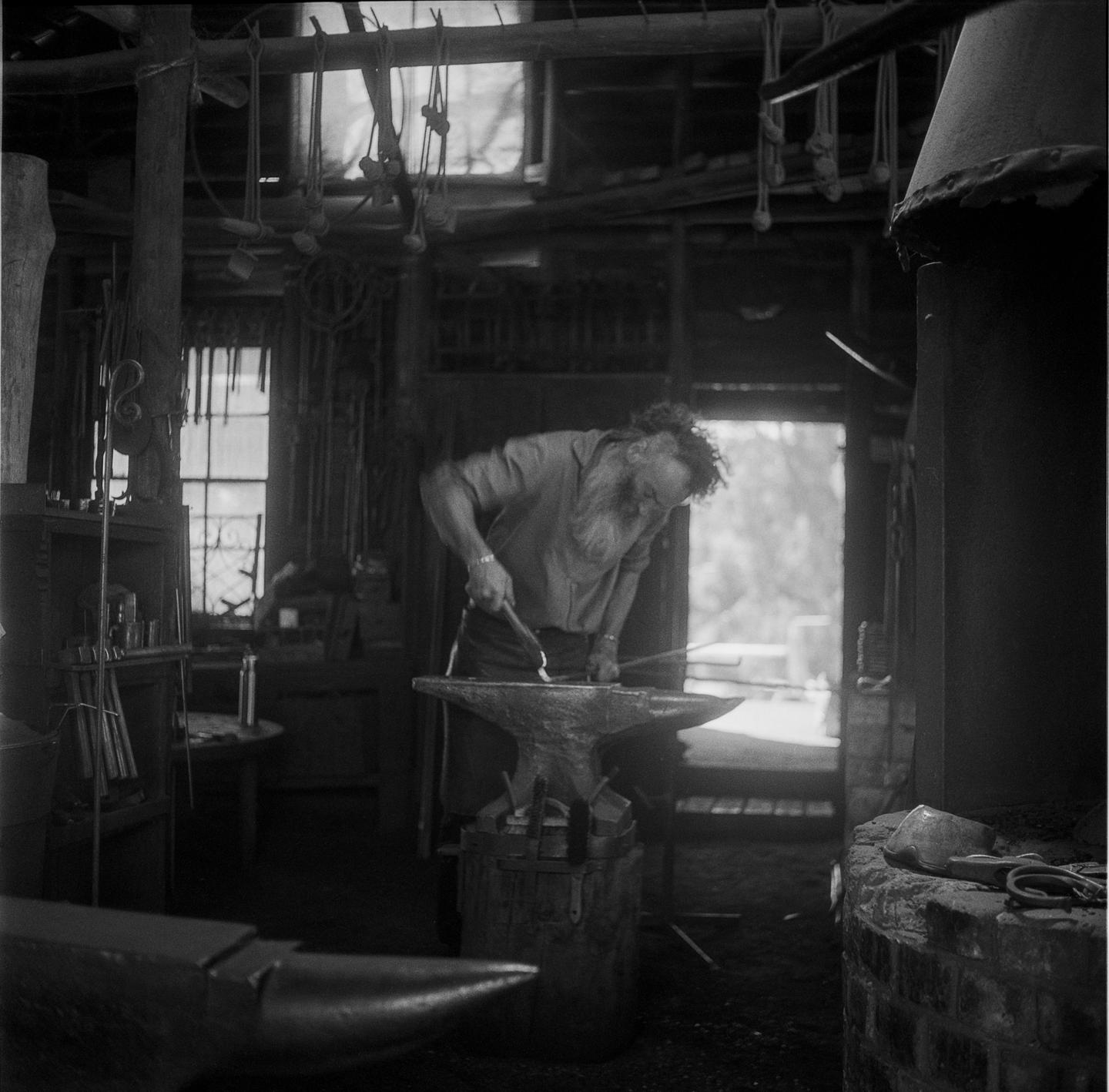 A man with a long beard works at an anvil in a blacksmith shop.