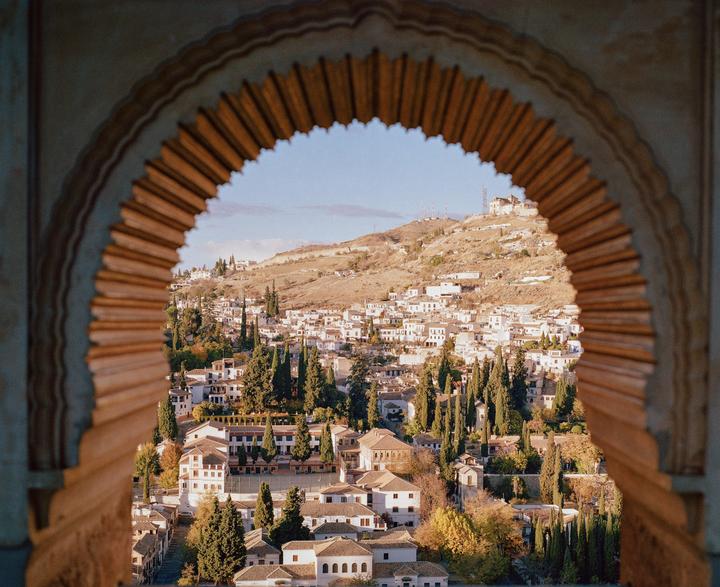 A view of a town with white buildings and cypress trees on a hillside, framed by an ornate archway.