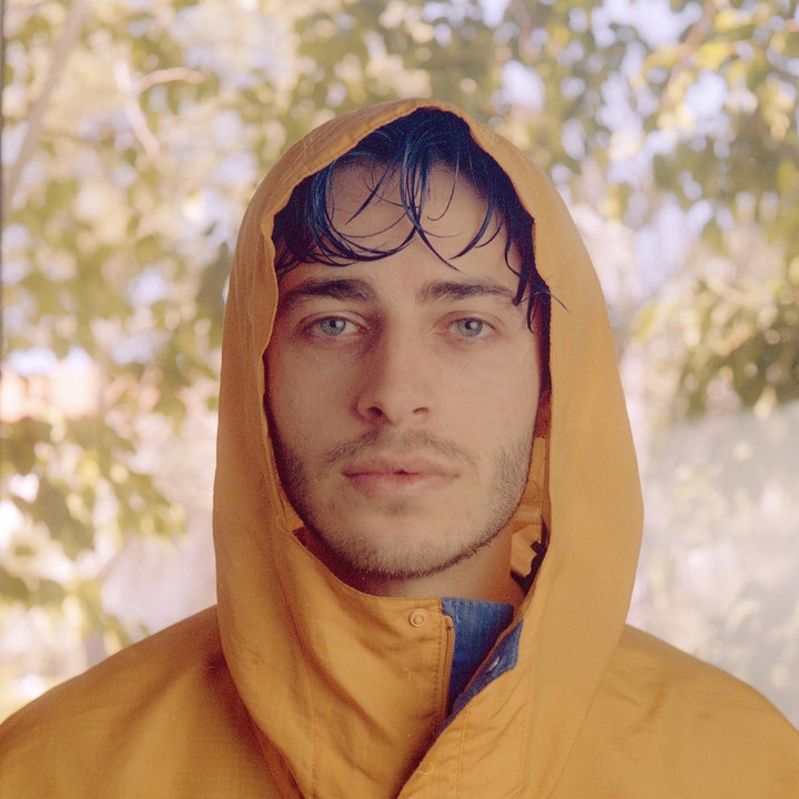 A man with wet hair looks directly at the viewer while wearing a yellow hooded jacket.