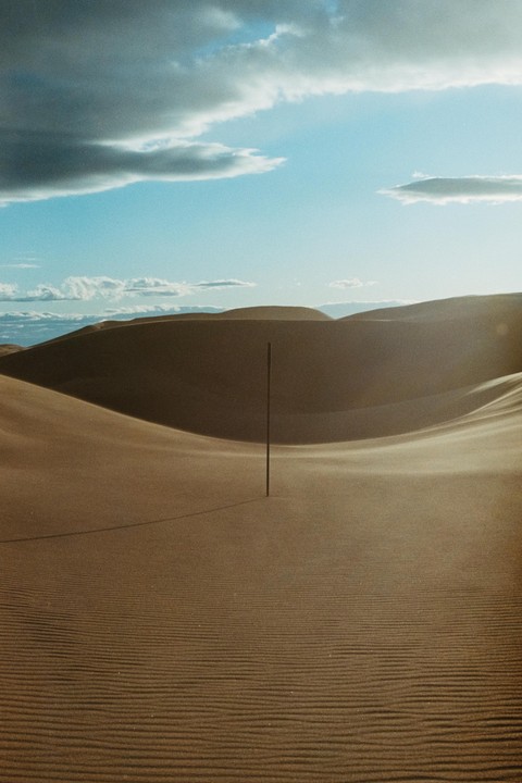 A stick stands upright in a vast desert landscape with sand dunes under a partly cloudy sky.