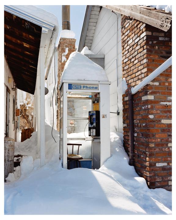 A white telephone booth is surrounded by deep snow between two buildings.