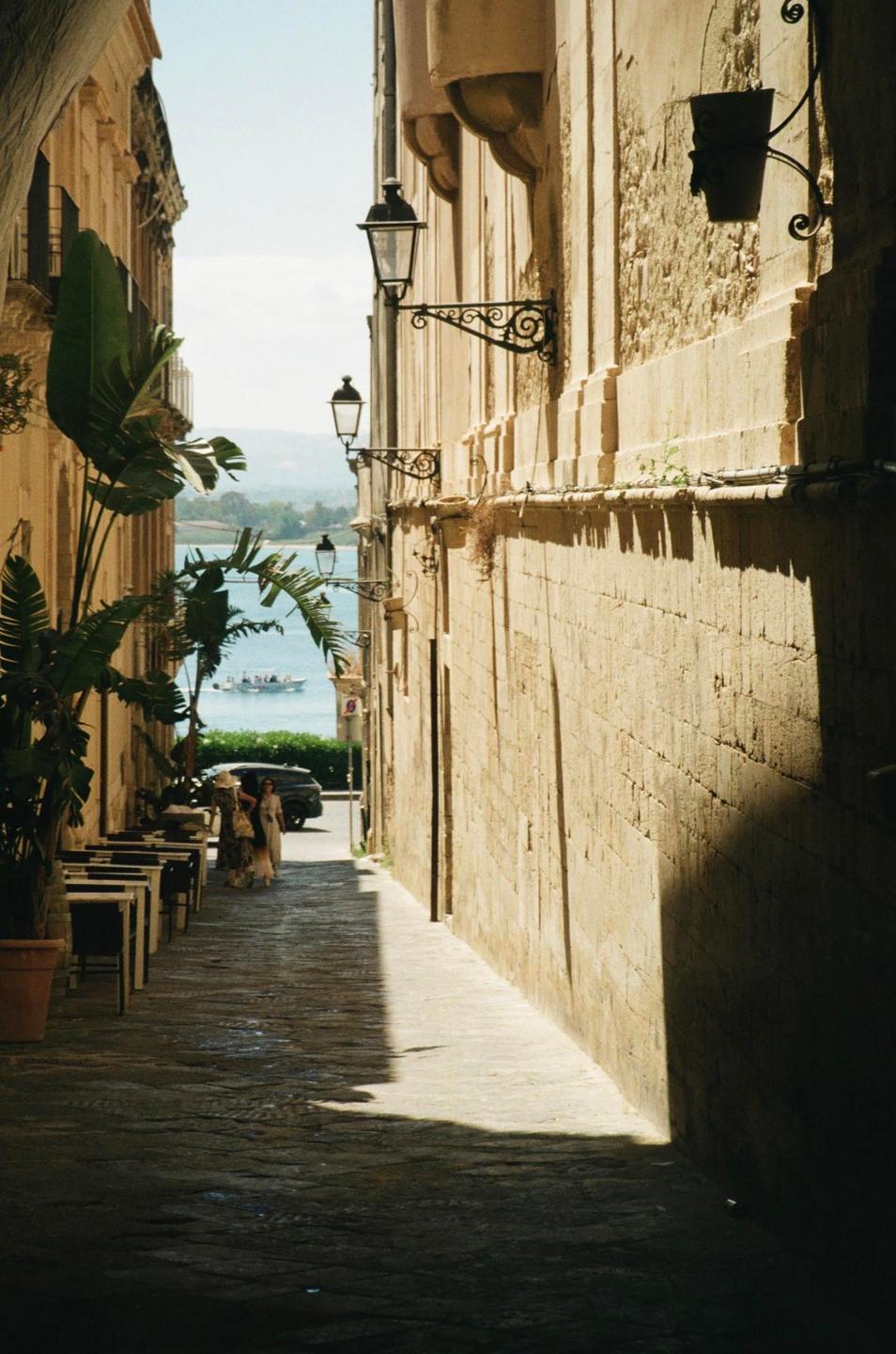 A narrow street with stone buildings and streetlights leads to a body of water with a boat.