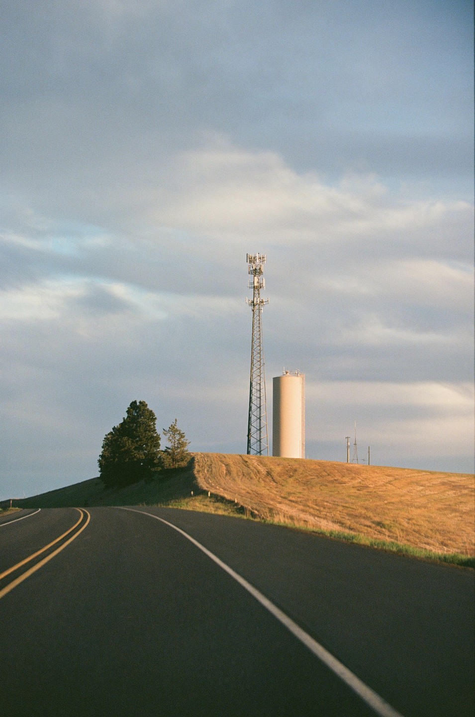 A road curves past a hill with a cell tower and water tower under a cloudy sky.