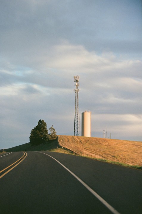 A road curves past a hill with a cell tower and water tower under a cloudy sky.