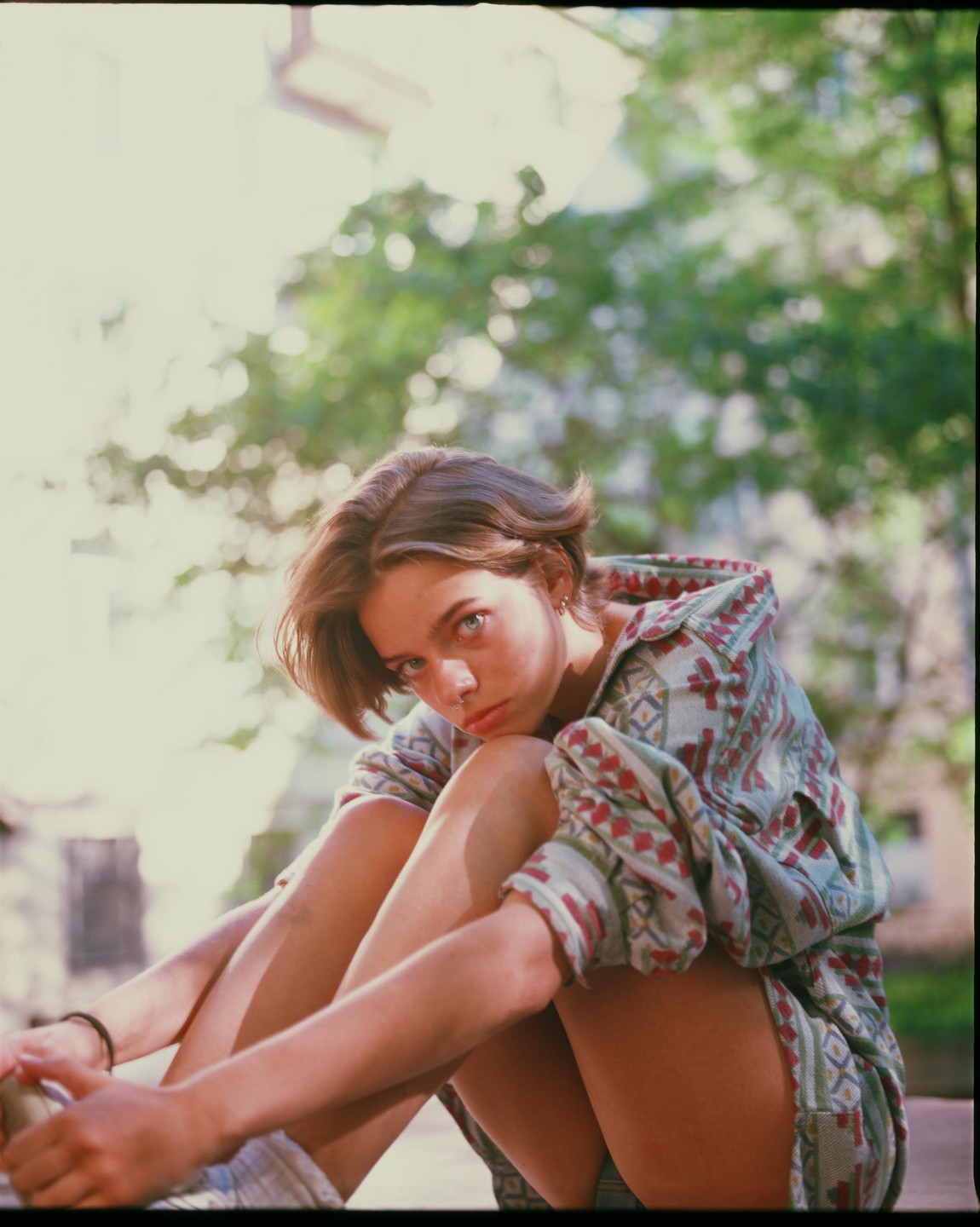 A young woman with short hair and a nose ring sits outdoors, looking directly at the viewer.