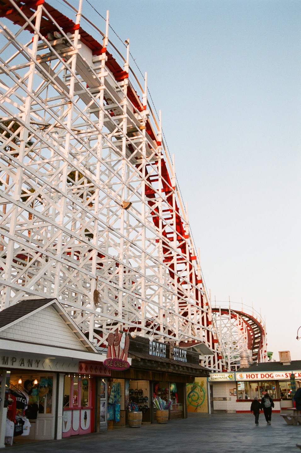 A large white and red rollercoaster dominates the frame above several small shops and a few people walking on a boardwalk.