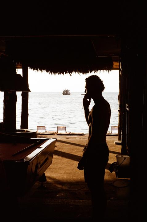 A man smokes a cigarette while standing next to a pool table with the ocean and a boat in the background.