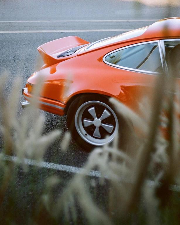 An orange sports car is parked on an asphalt road with a white line, partially obscured by tall grass in the foreground.