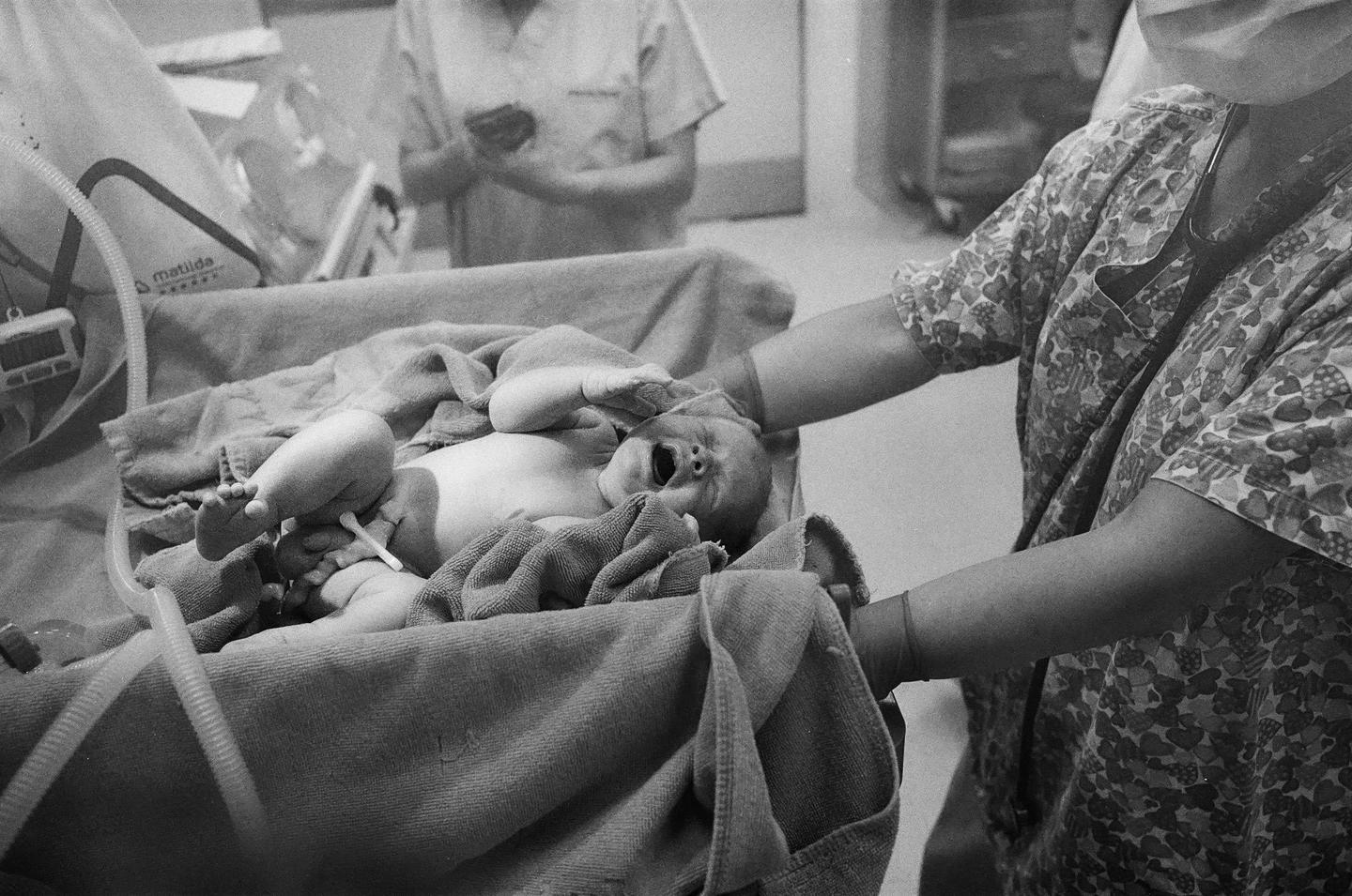 A newborn baby lies on a blanket with medical equipment nearby, while a person in scrubs tends to the baby.