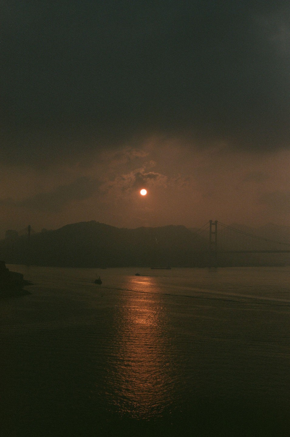 The sun sets over a body of water with a bridge and hills in the distance.