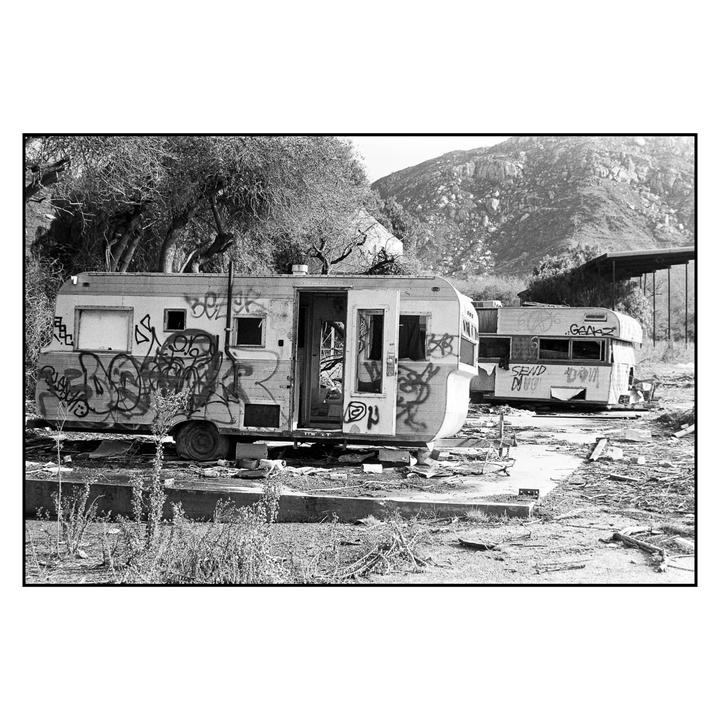 Two abandoned and vandalized recreational vehicles sit in a desolate, overgrown area with a mountain in the background.